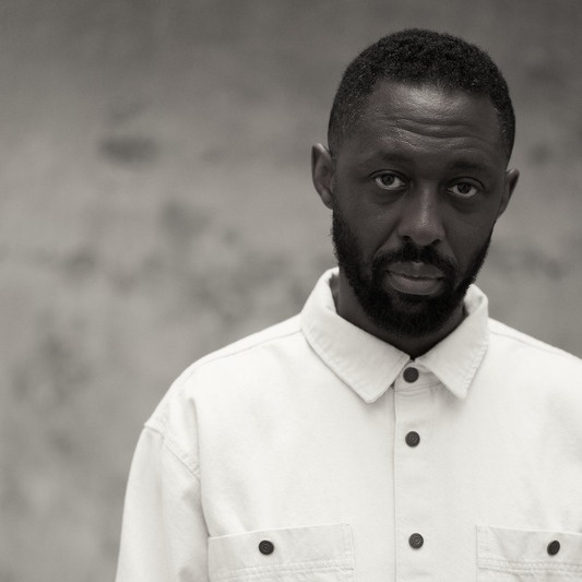 Visuel en noir et blanc de l'acteur et humoriste Thomas Ngijol, ambassadeur officiel du NollywoodWeek Film Festival 2026, portant une chemise en jean blanche et regardant droit face à la caméra.